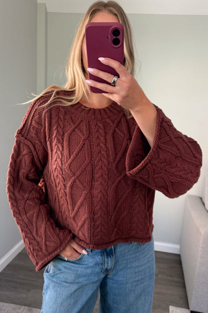 A woman takes a mirror selfie wearing a rust-brown cable-knit cropped sweater and light blue jeans in a home setting.