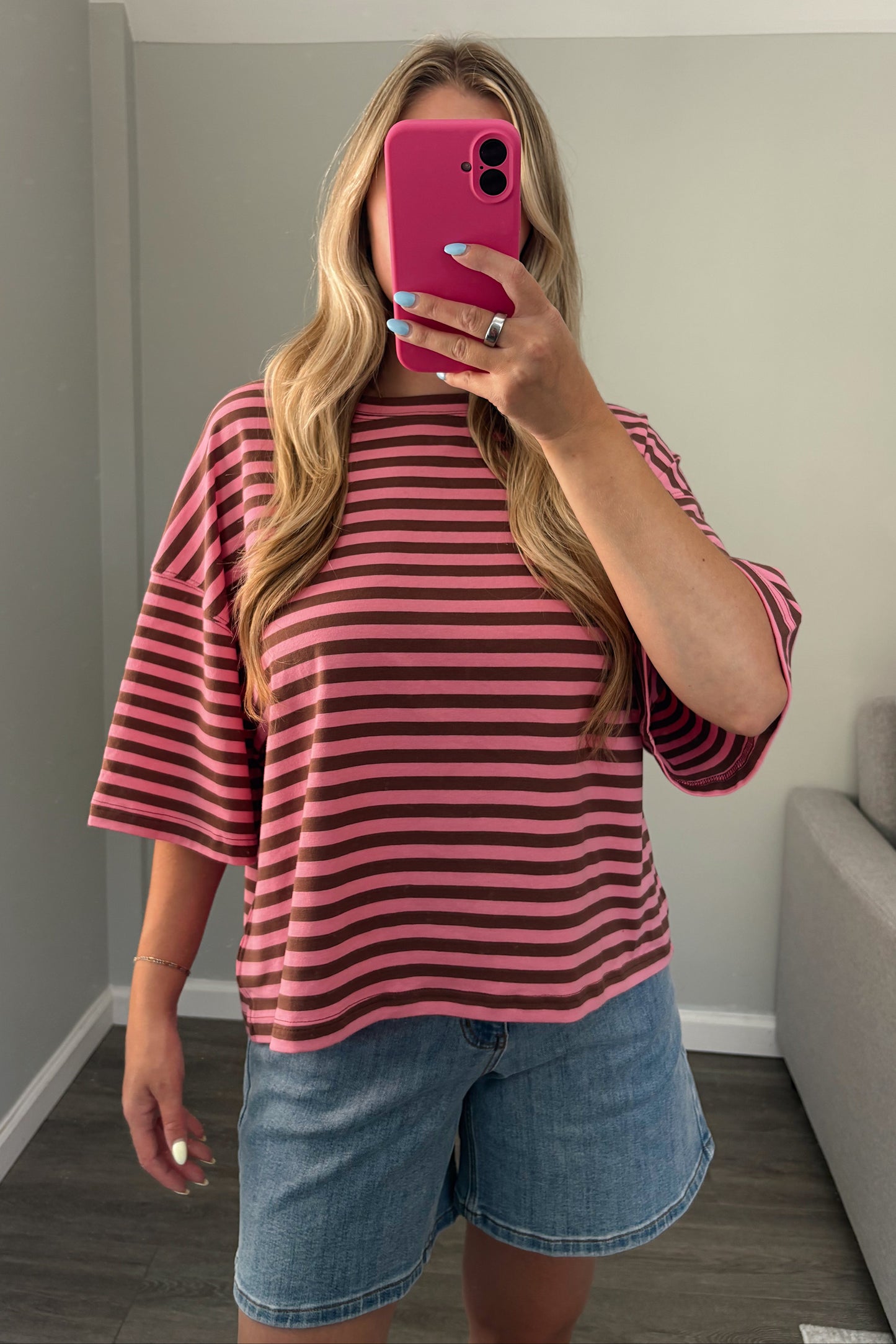 A woman takes a mirror selfie wearing a pink and dark brown striped short-sleeve t-shirt and denim shorts in a home setting.