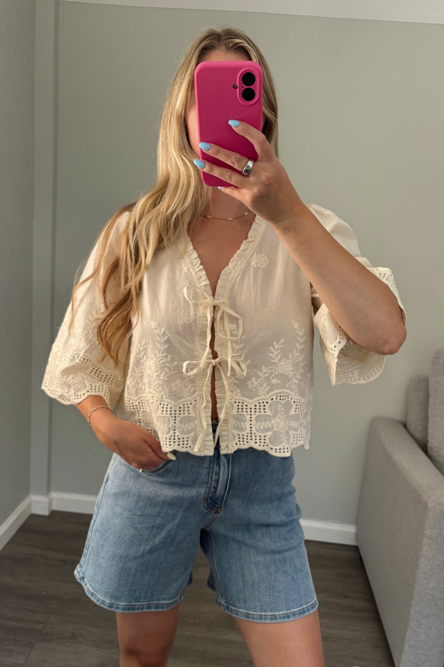 Woman taking a mirror selfie wearing a white lace top and denim shorts in a room with a gray couch.