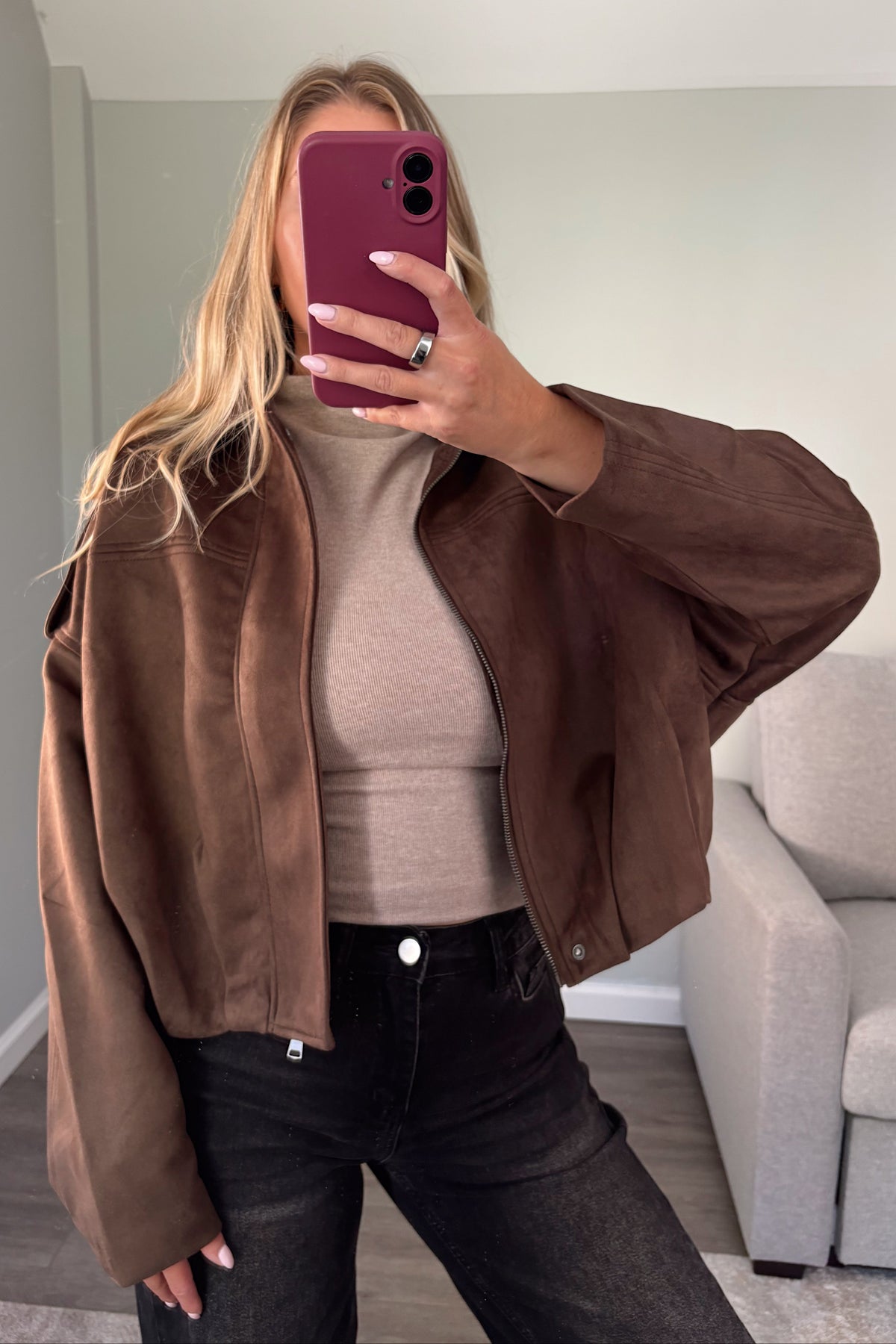 A woman takes a mirror selfie wearing a brown suede-like cropped jacket over a beige ribbed top and black jeans, holding a burgundy smartphone.