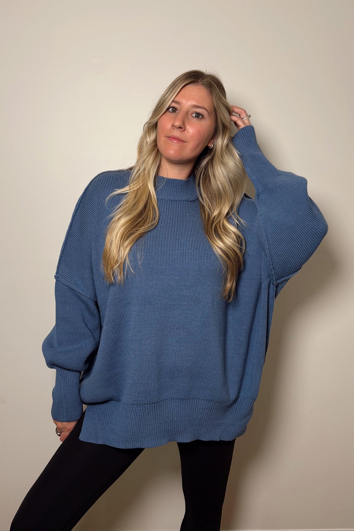 Front view of the Essential dusty blue ribbed knit sweater with one arm raised, showing its roomy sleeves and fitted cuffs.

