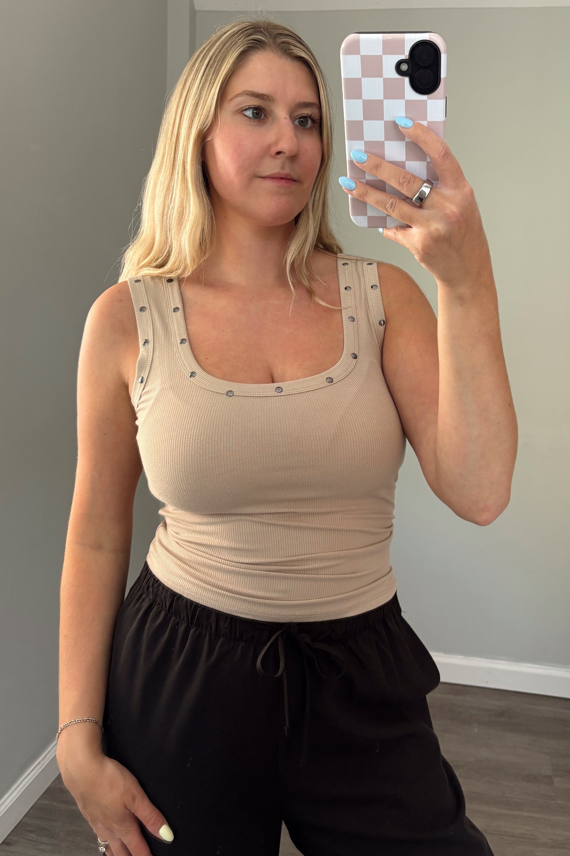 Woman taking a mirror selfie wearing a beige top and black pants in a room with a gray couch.