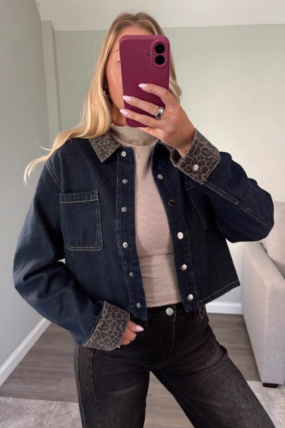A woman takes a mirror selfie wearing a cropped dark blue denim jacket with leopard-print collar and cuff accents, a beige ribbed top, and dark jeans.