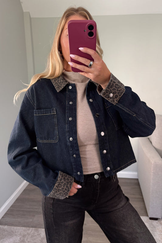 A woman takes a mirror selfie wearing a cropped dark blue denim jacket with leopard-print collar and cuff accents, a beige ribbed top, and dark jeans.