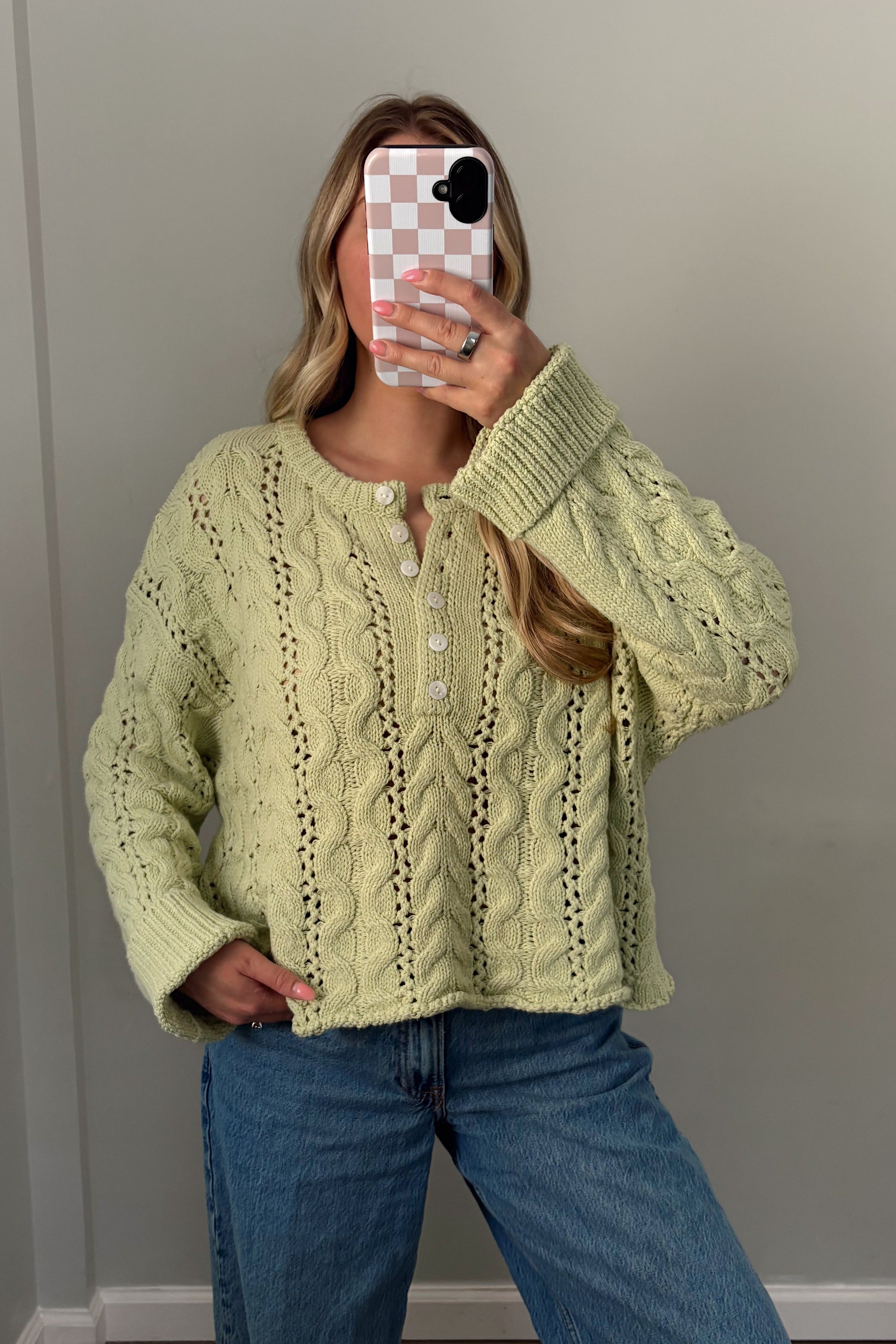 Person wearing a light green cable knit sweater and blue jeans, standing indoors.