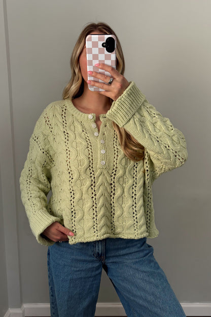 Person wearing a light green cable knit sweater and blue jeans, standing indoors.