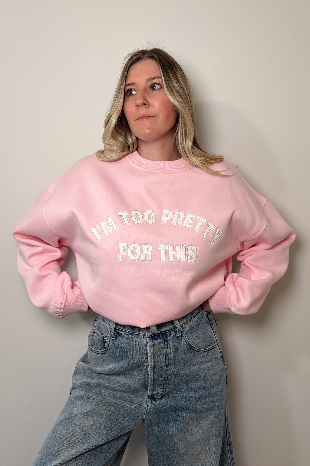 Too Pretty pink crewneck sweatshirt tucked into high-rise blue jeans, showing the white embroidered slogan across the chest.

