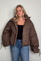 Model in the Coco leopard print puffer jacket worn open over a black top, with drawstring ties at the sleeve cuffs and blue jeans