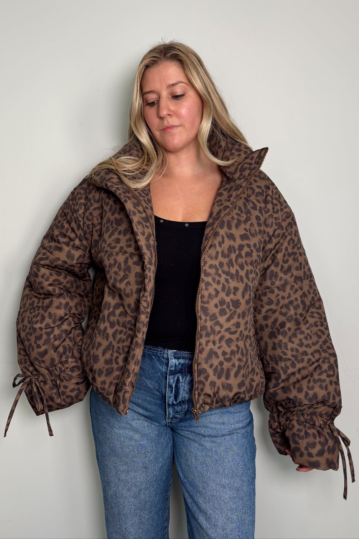 Model in the Coco leopard print puffer jacket worn open over a black top, with drawstring ties at the sleeve cuffs and blue jeans