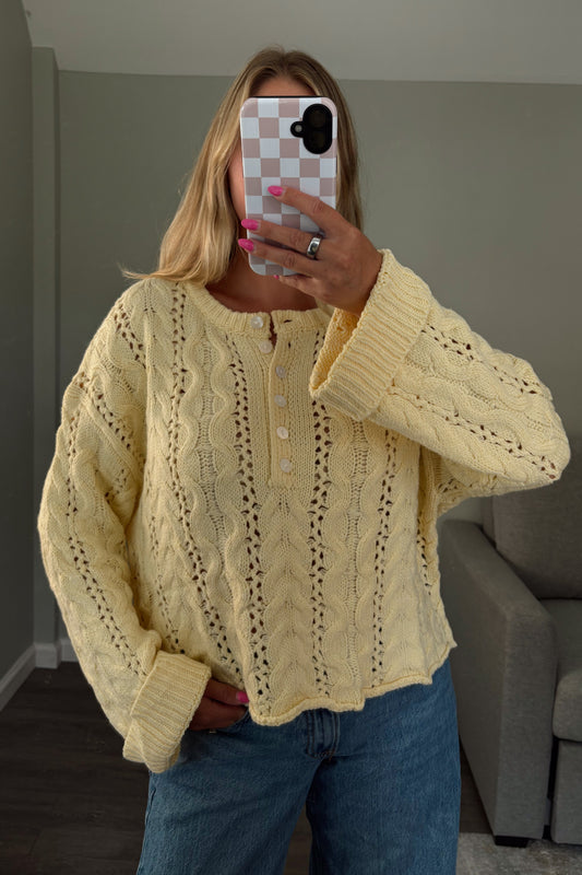 A woman takes a mirror selfie wearing a pale yellow cable-knit button-up sweater and blue jeans in a room with gray walls and a gray sofa.