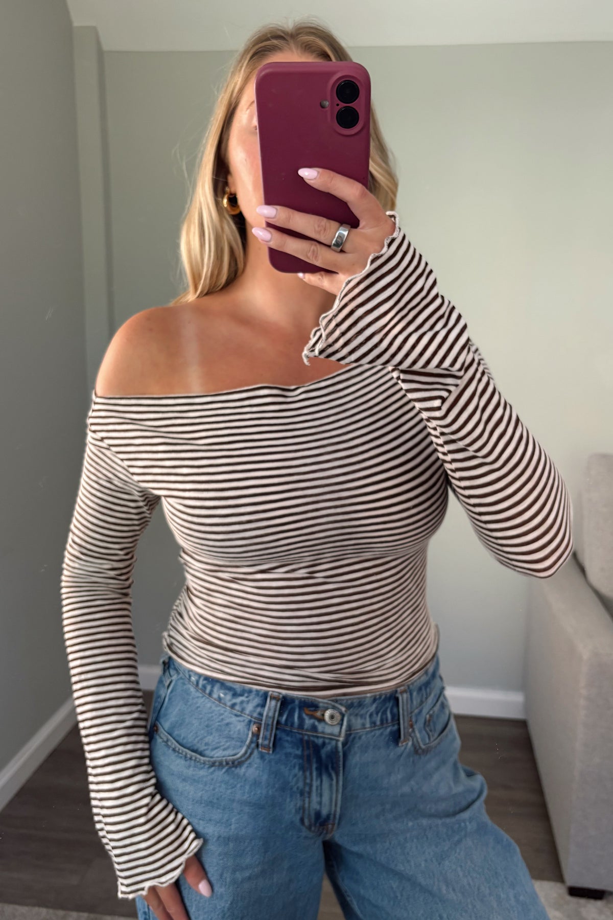 A woman takes a mirror selfie wearing a beige and black horizontally striped off-the-shoulder long-sleeve top with flared cuffs, paired with blue jeans.