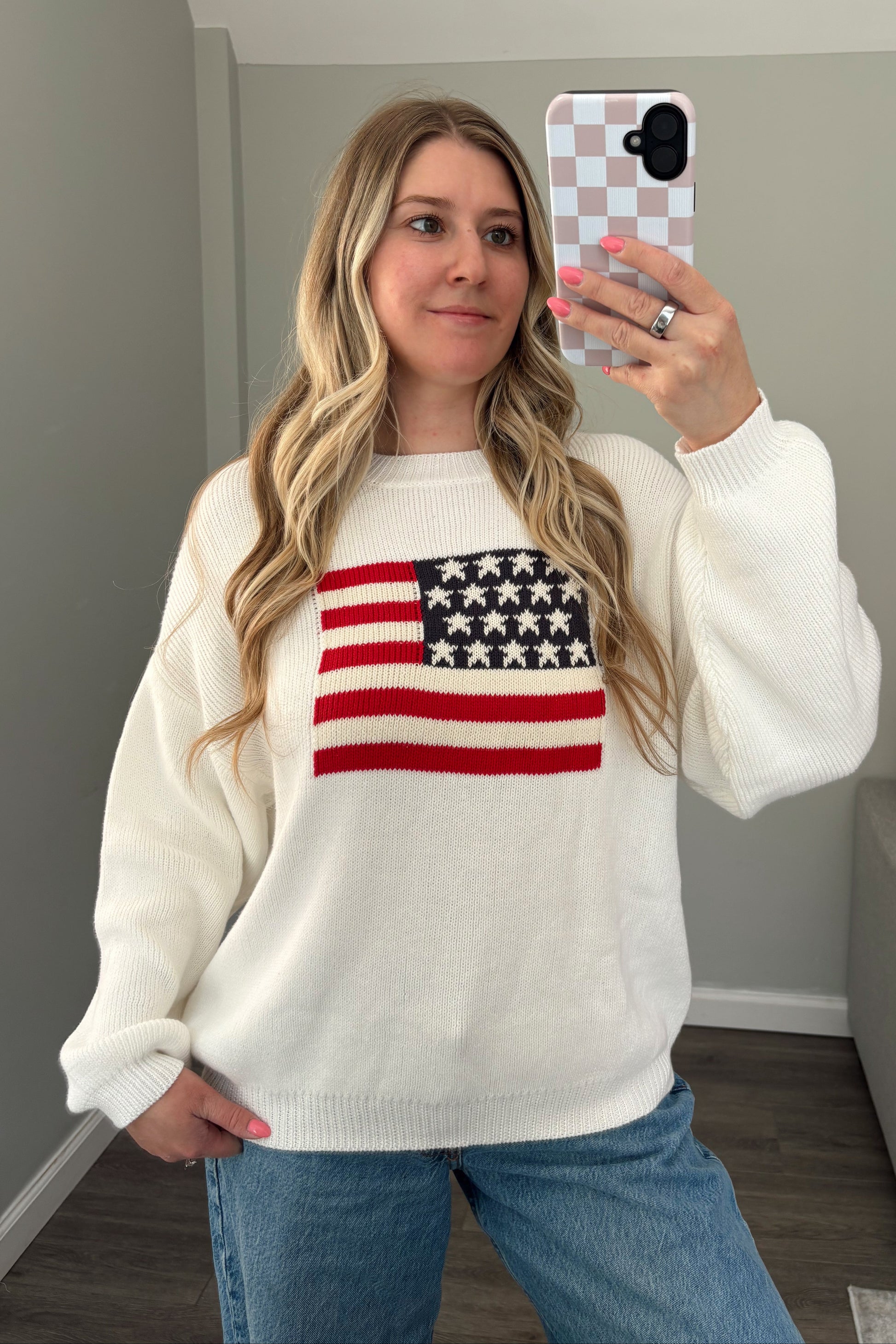 Person wearing a white sweater with an American flag design, taking a mirror selfie.