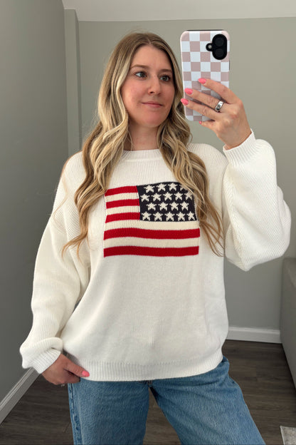 Person wearing a white sweater with an American flag design, taking a mirror selfie.