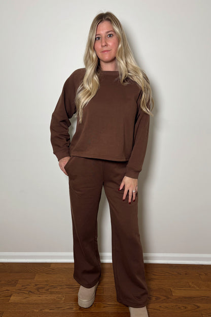 Woman wearing a brown sweat set/lounge set standing against a white wall.