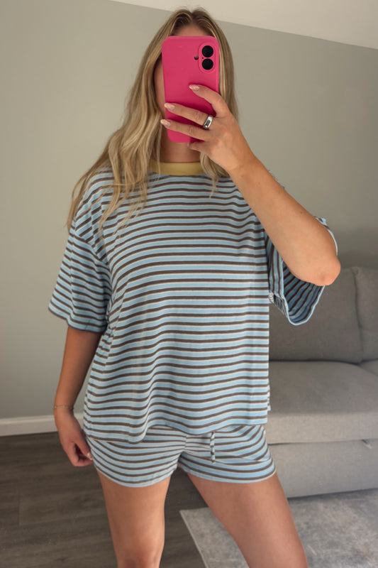 A woman takes a mirror selfie wearing a matching two-piece loungewear set consisting of a short-sleeve striped t-shirt and shorts in light blue and white. She is holding a bright pink smartphone in front of her face, with a gray sofa and wooden floor visible in the background.