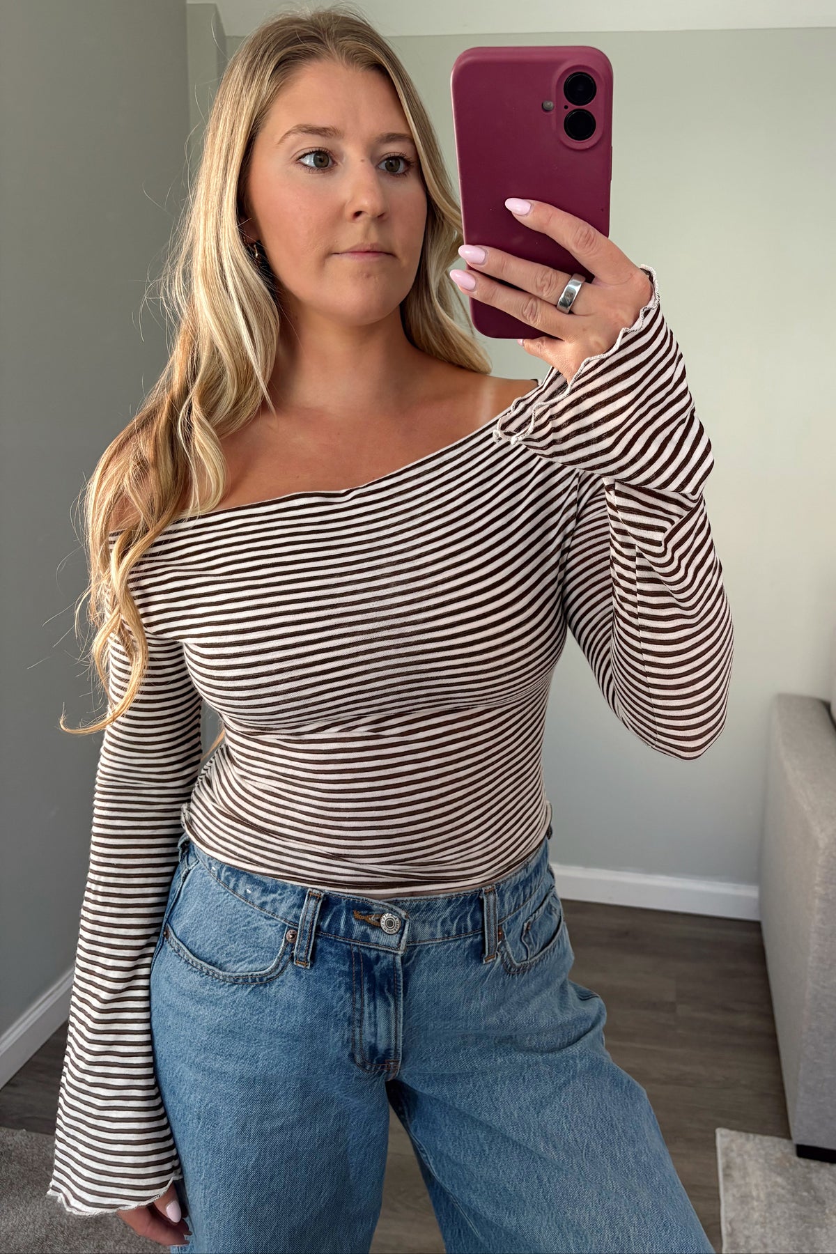 A woman taking a mirror selfie wearing a brown and white horizontally striped off-the-shoulder long-sleeve top and blue jeans.