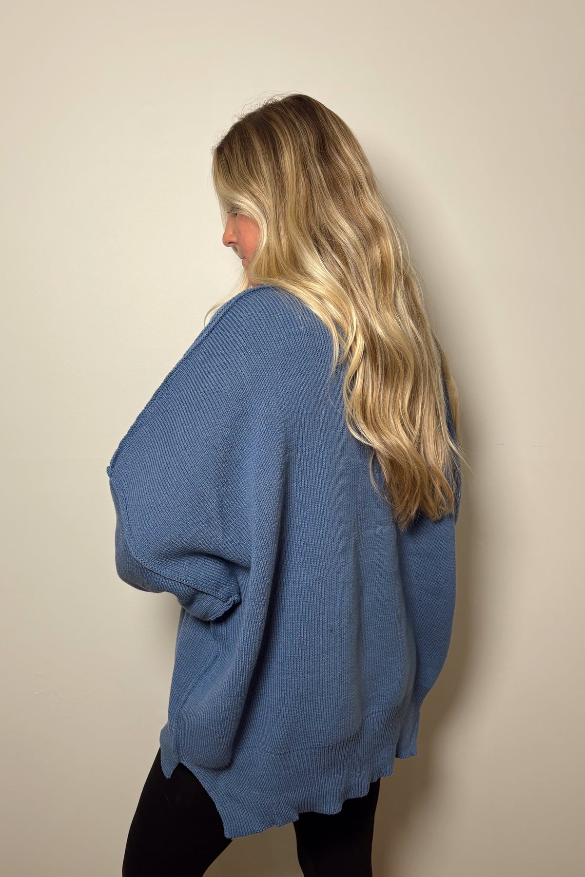Side and back view of the Essential dusty blue knit sweater showing its loose drape, ribbed hem, and side slit.

