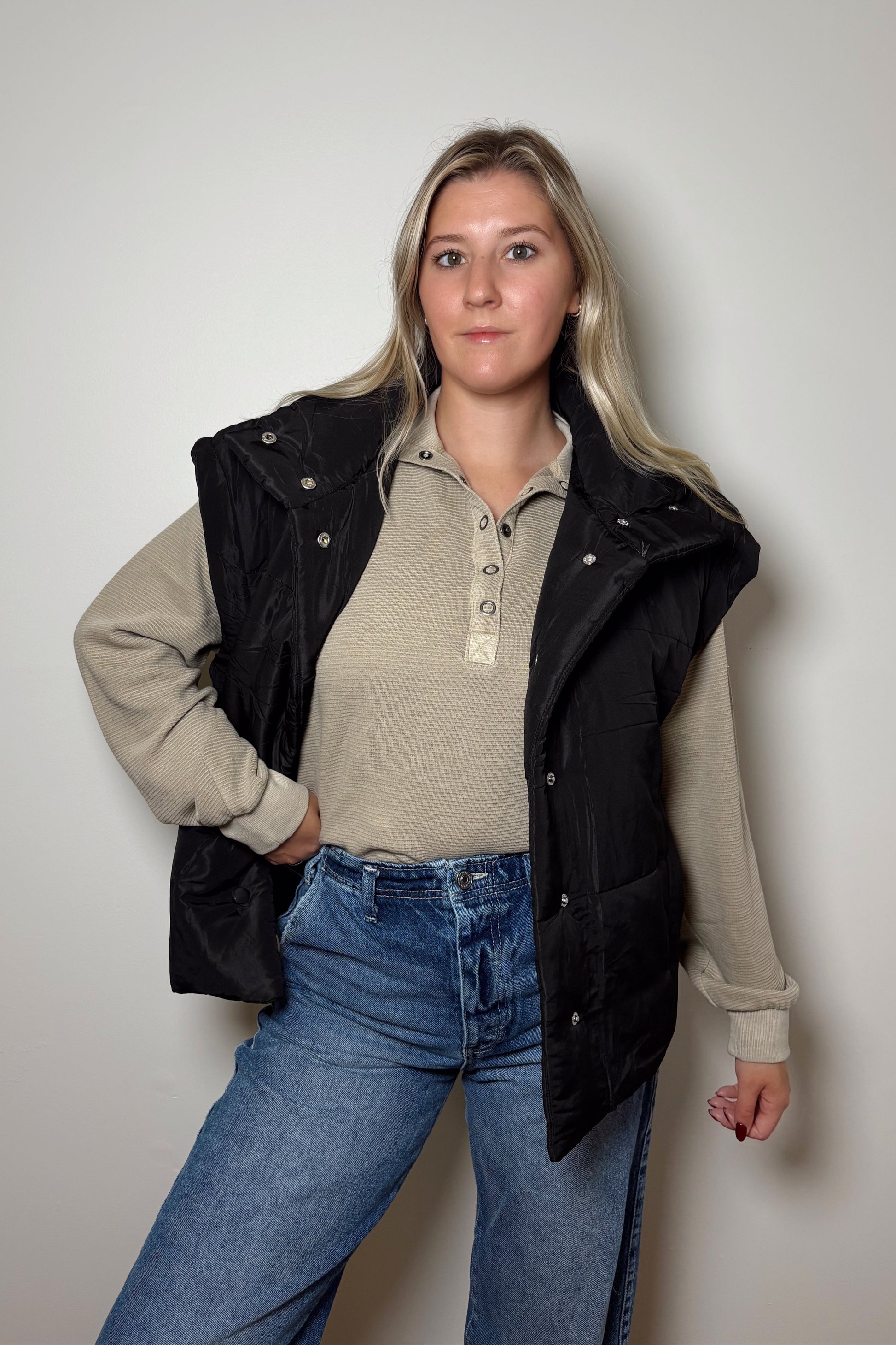Woman wearing a beige shirt, black vest, and blue jeans against a plain background