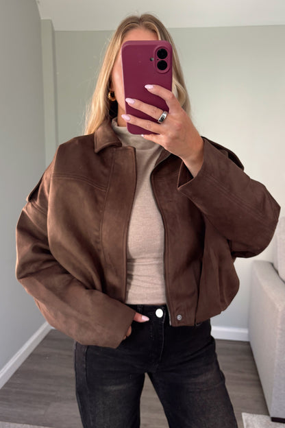 A woman takes a mirror selfie wearing a brown cropped suede jacket over a beige turtleneck and black jeans, holding a dark magenta smartphone.