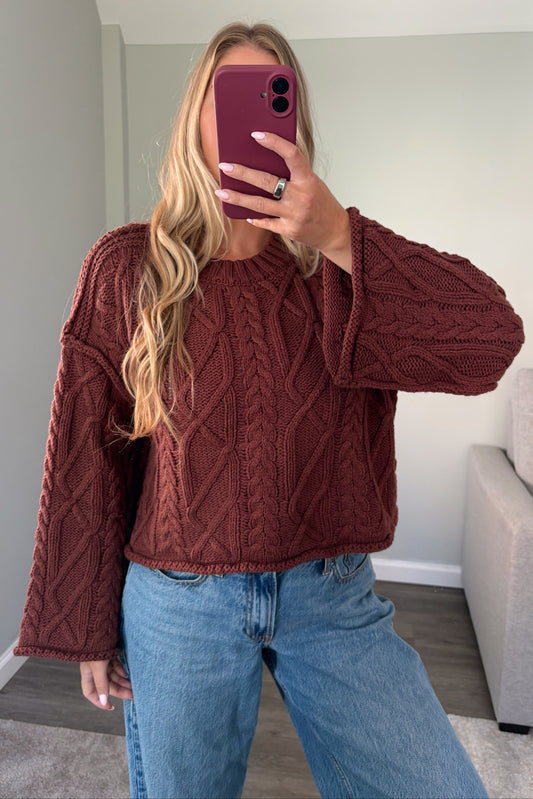 A woman takes a mirror selfie wearing a rust-brown cable-knit cropped sweater and light blue jeans in a home setting.