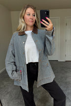 Derby striped denim barn jacket worn open over a white tee and black jeans, with one hand in a front pocket and the brown corduroy collar visible.

