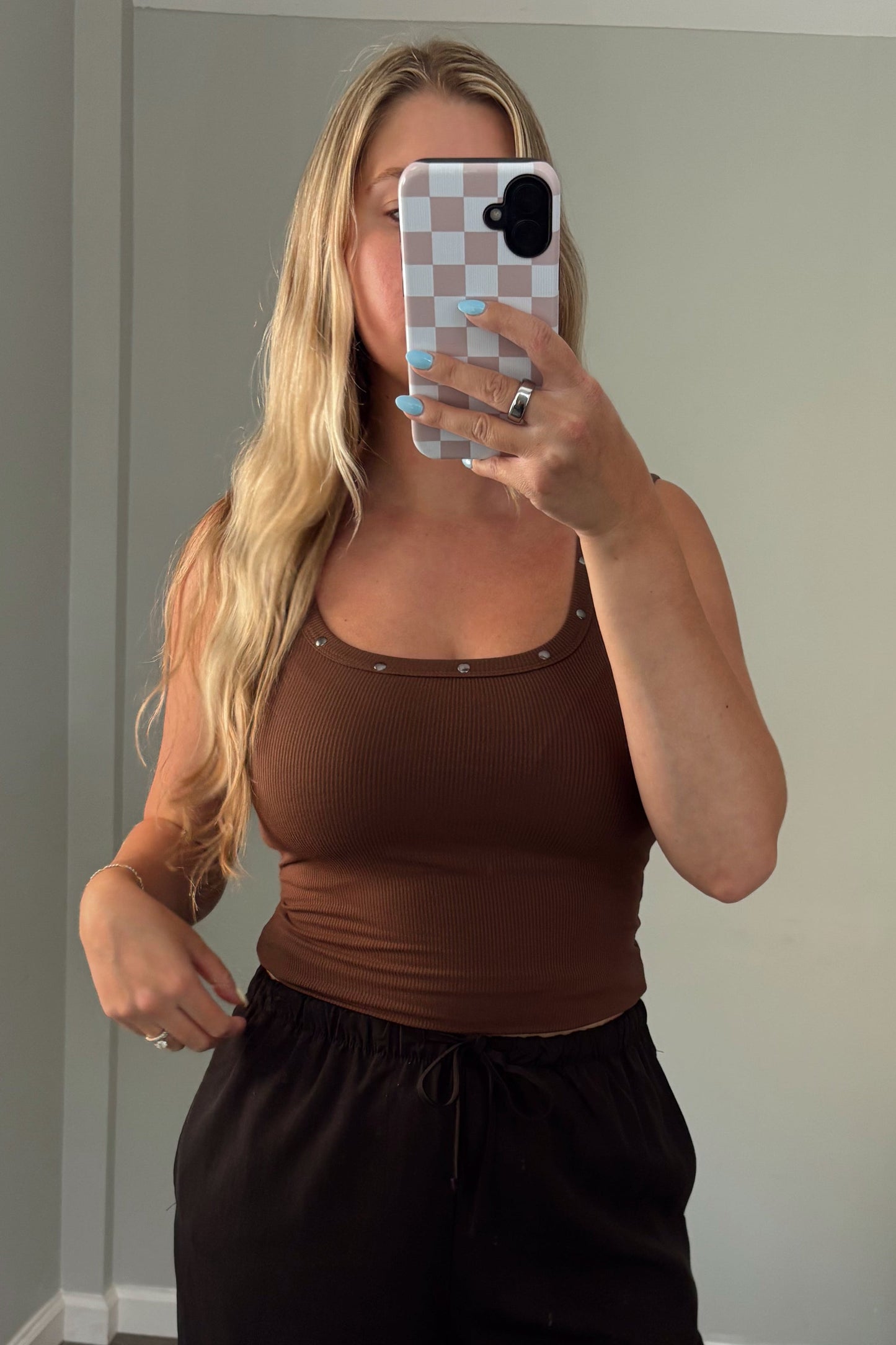 Person wearing a brown top and black pants taking a mirror selfie in a room with a couch and window.