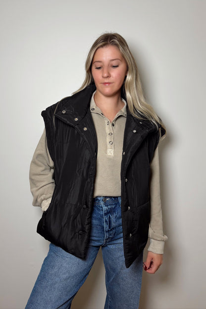 Person wearing a black vest over a beige jacket with a plain background