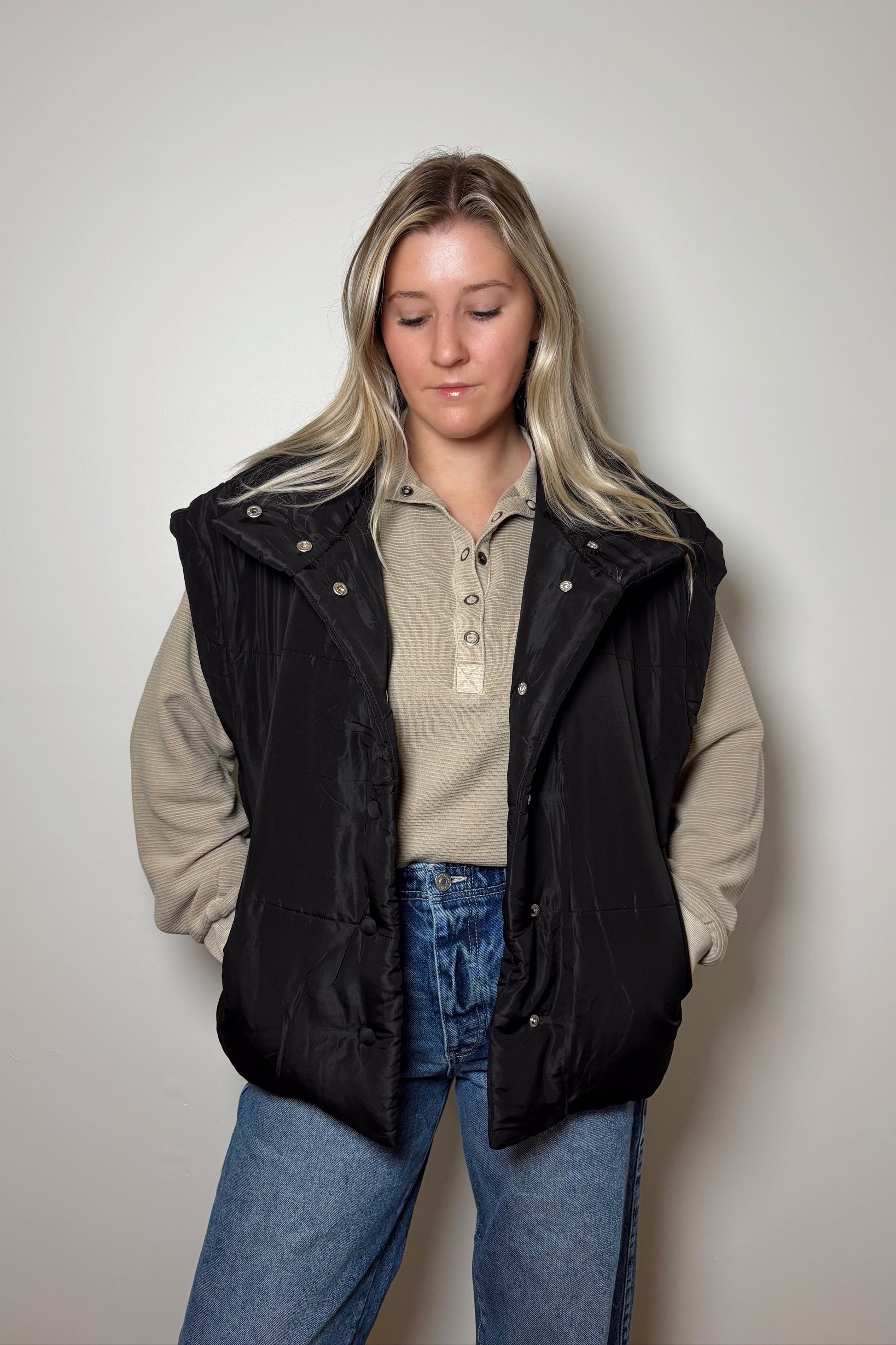 Person wearing a black puffer vest over a beige shirt and blue jeans on a plain background