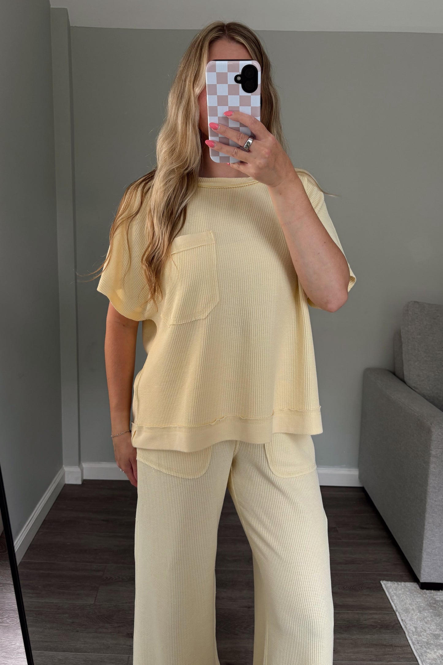 Person wearing a light yellow outfit taking a mirror selfie in a room with a gray sofa.