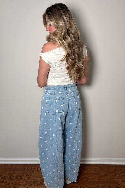 Woman wearing a white top and light blue polka dot pants against a plain wall.