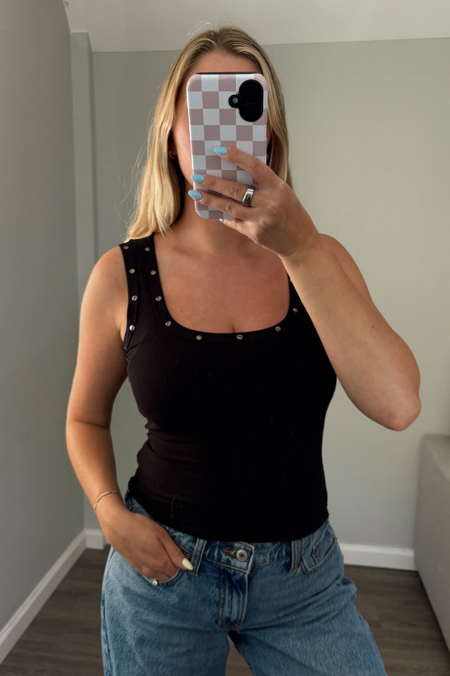 Person wearing a black tank top with studs and blue jeans, taking a mirror selfie.