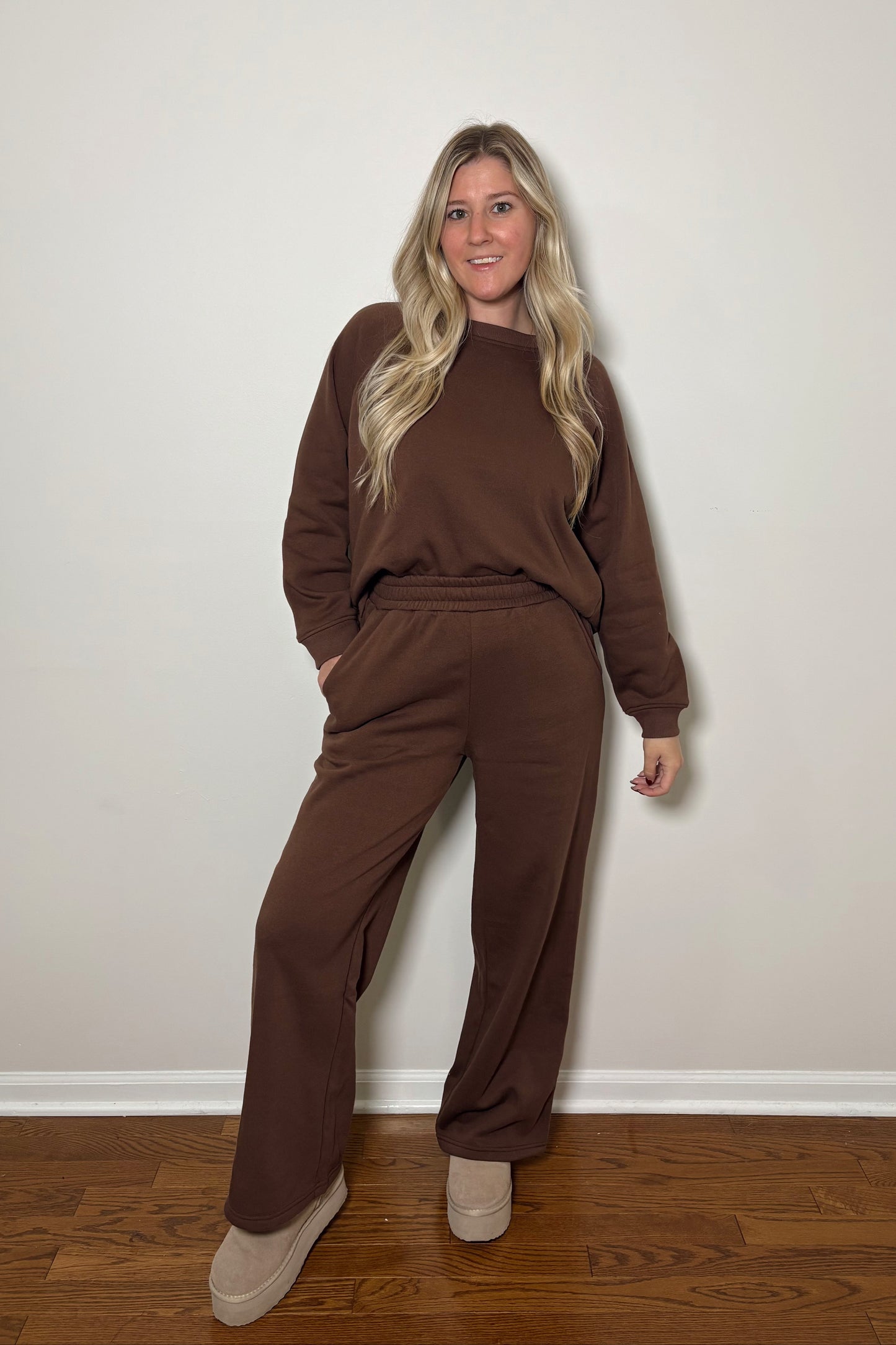 Woman wearing a brown sweat set/lounge set standing against a plain wall.