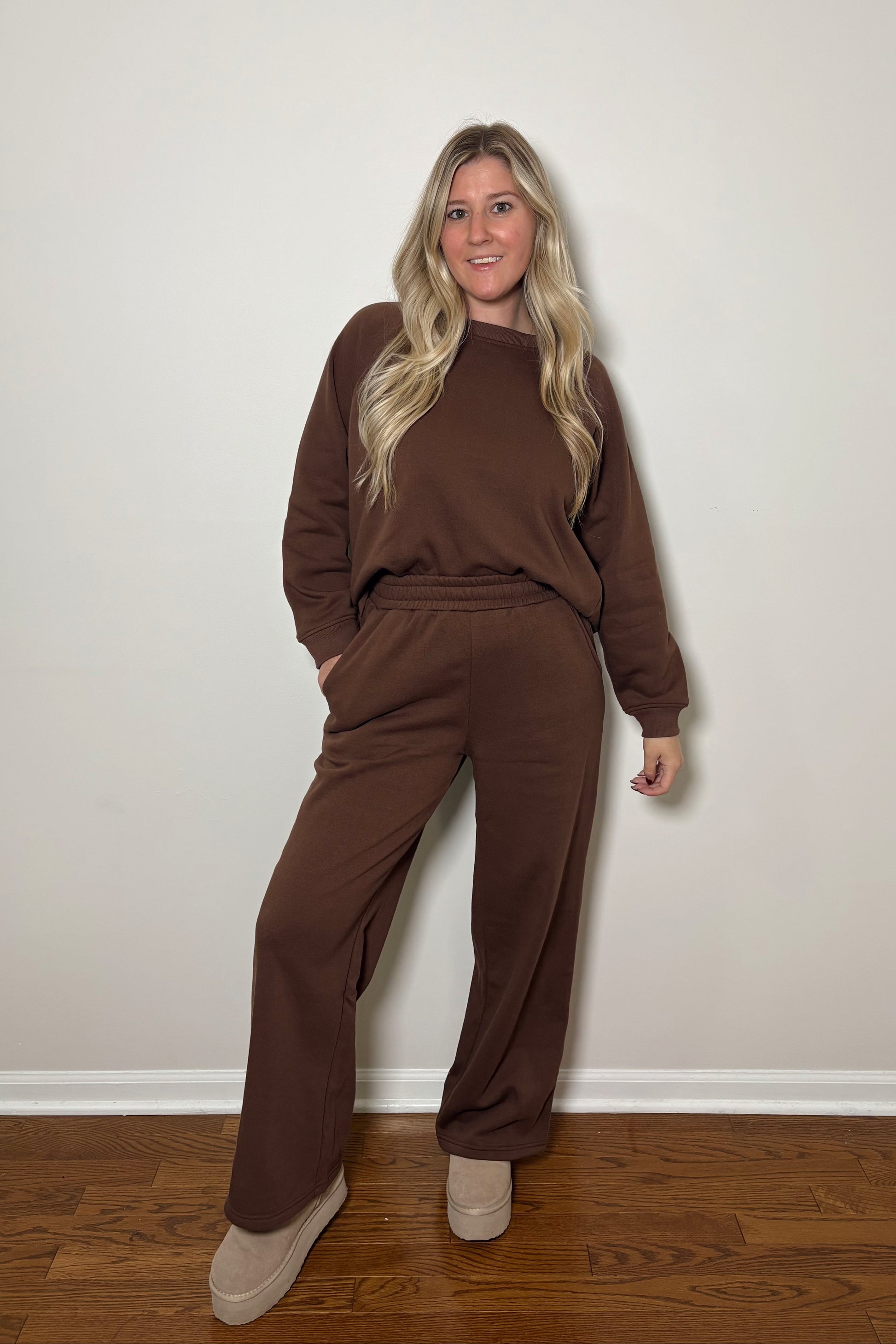 Woman wearing a brown sweat set/lounge set standing against a plain wall.
