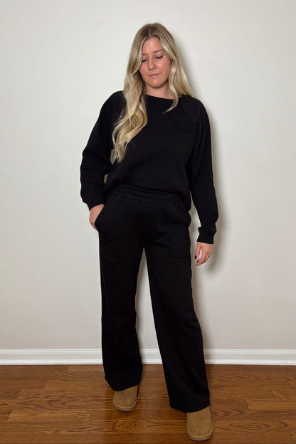 Person wearing a black lounge set standing against a plain white wall.