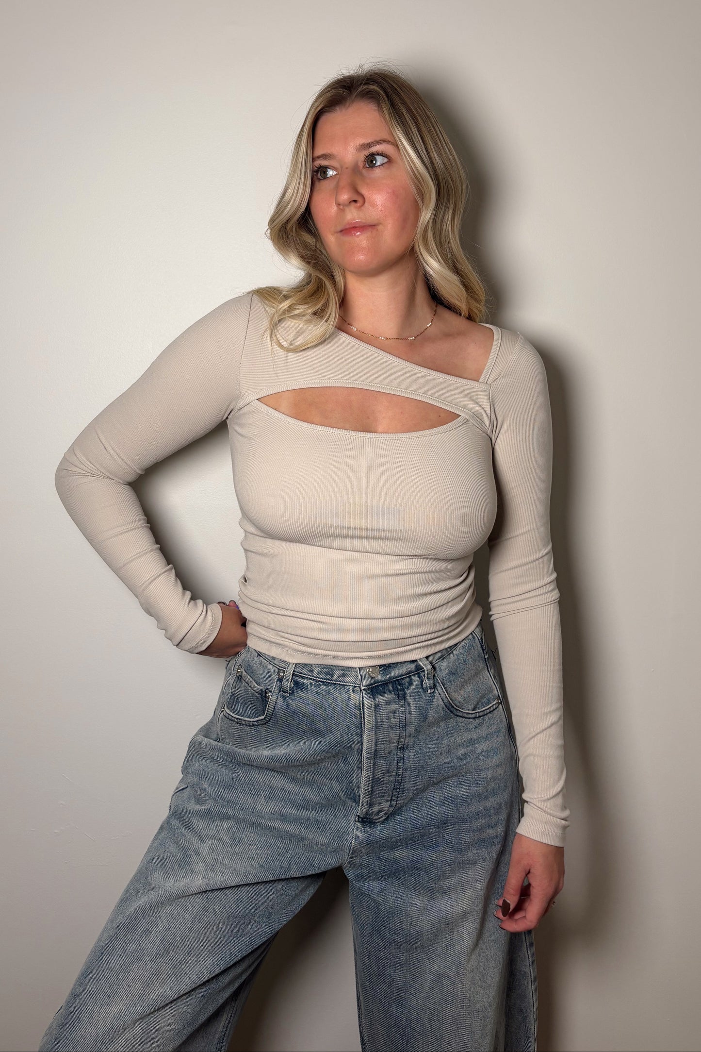 Person wearing a beige long-sleeve top with cut-out design and blue jeans against a plain background