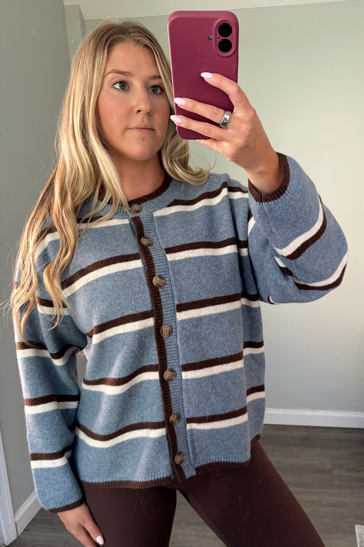 A woman takes a mirror selfie wearing a blue knit cardigan with brown and cream horizontal stripes, paired with dark brown leggings in a home setting.