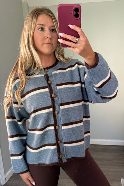 A woman takes a mirror selfie wearing a blue knit cardigan with brown and cream horizontal stripes, paired with dark brown leggings in a home setting.