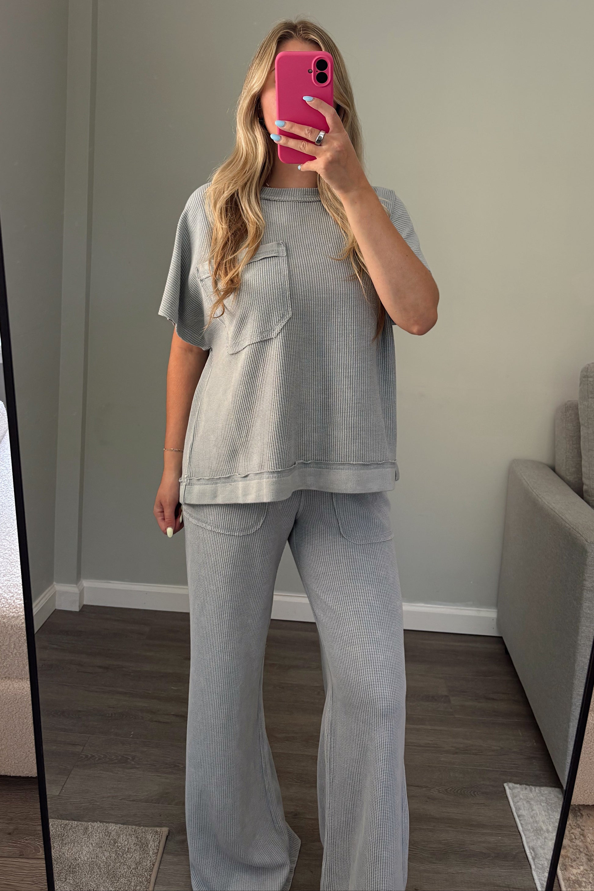 Person taking a mirror selfie wearing a blue t-shirt and light blue pants in a room with a couch and window.