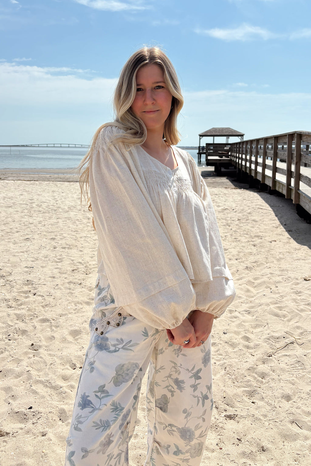 Woman wearing the Salt Air blouse with long bubble sleeves and floral pants beside a beach pier