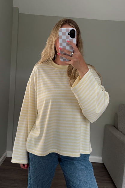 Person wearing a yellow and white striped shirt and blue jeans taking a mirror selfie in a room with a gray couch.