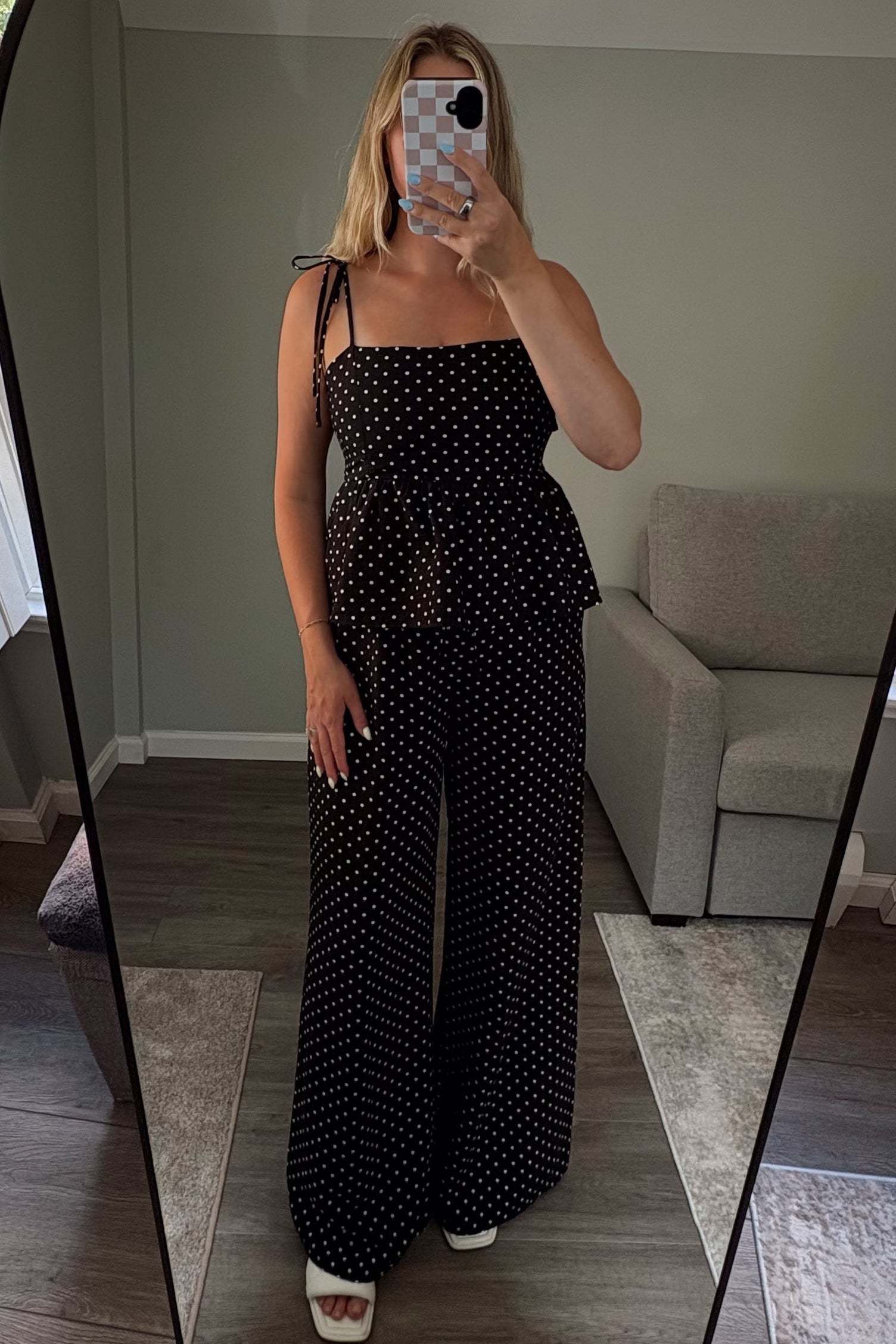 Person taking a mirror selfie wearing a black polka dot outfit in a room with large windows.