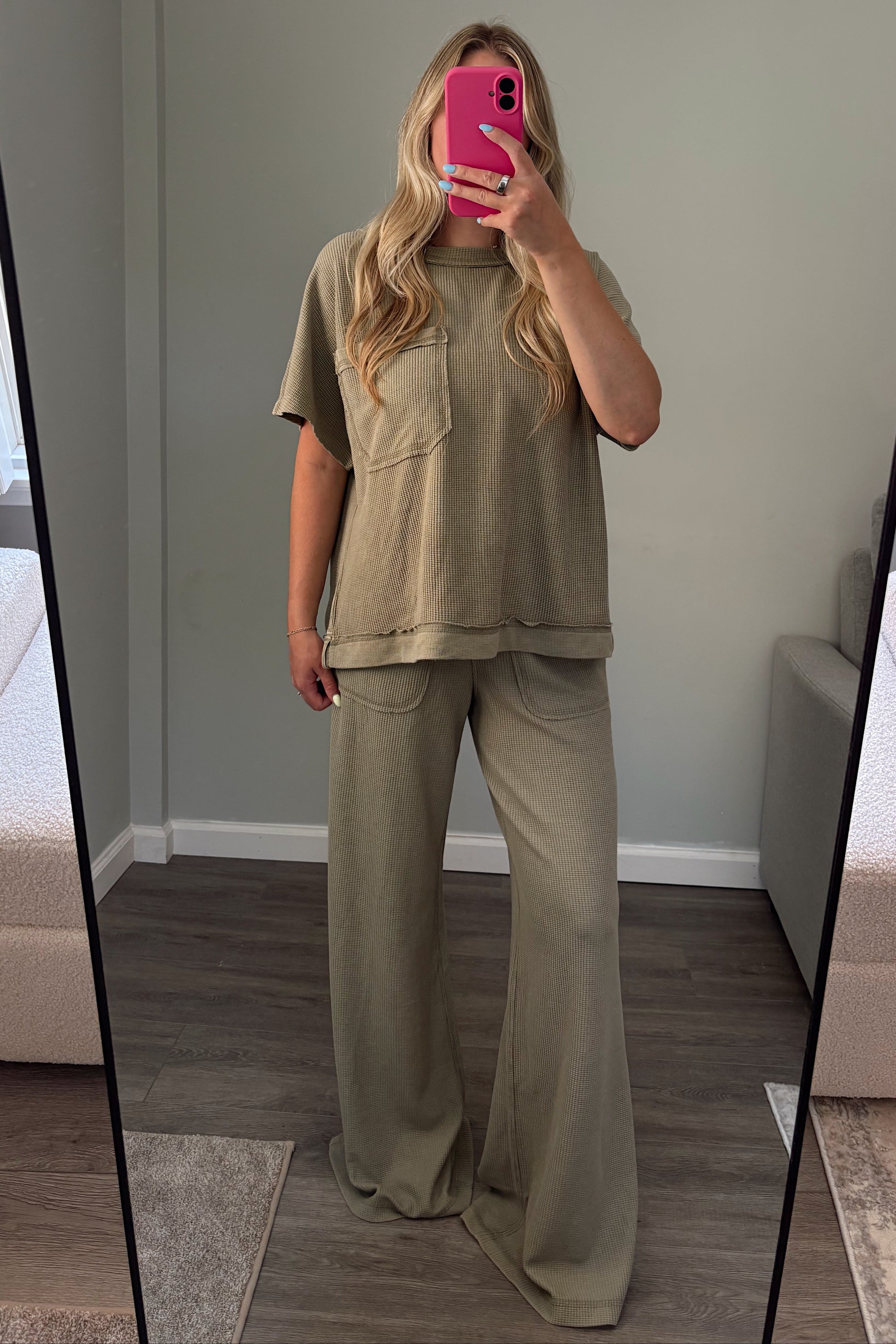 A woman takes a mirror selfie in a room wearing a matching two-piece lounge set consisting of a short-sleeve textured top and wide-leg pants in a muted sage green color.
