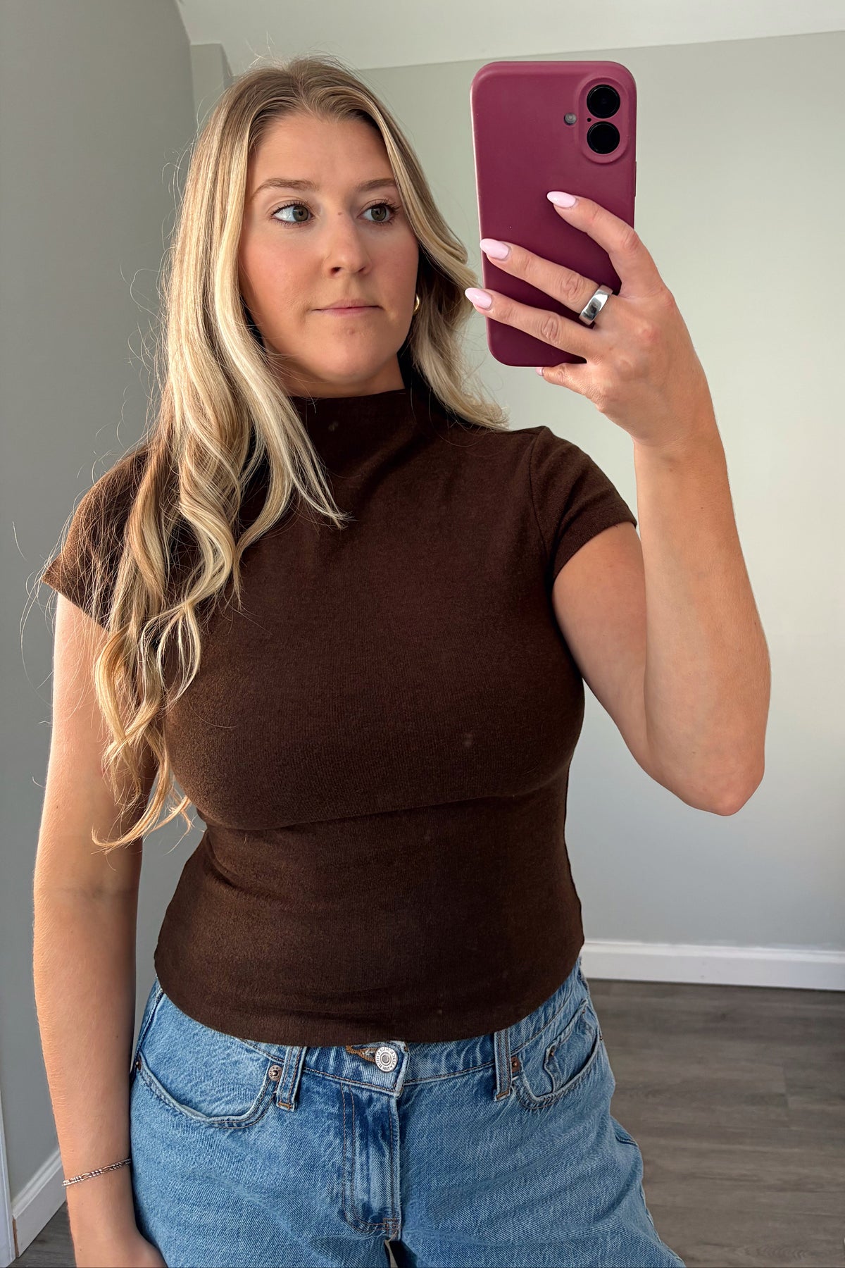 A woman taking a mirror selfie wearing a fitted short-sleeve brown mock neck top and blue jeans, holding a magenta phone.