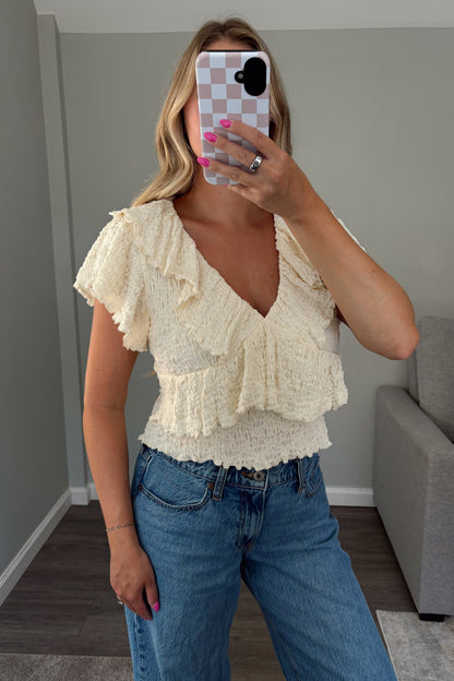 A woman takes a mirror selfie wearing a cream ruffled short-sleeve top and blue jeans in a room with gray walls and a gray armchair.