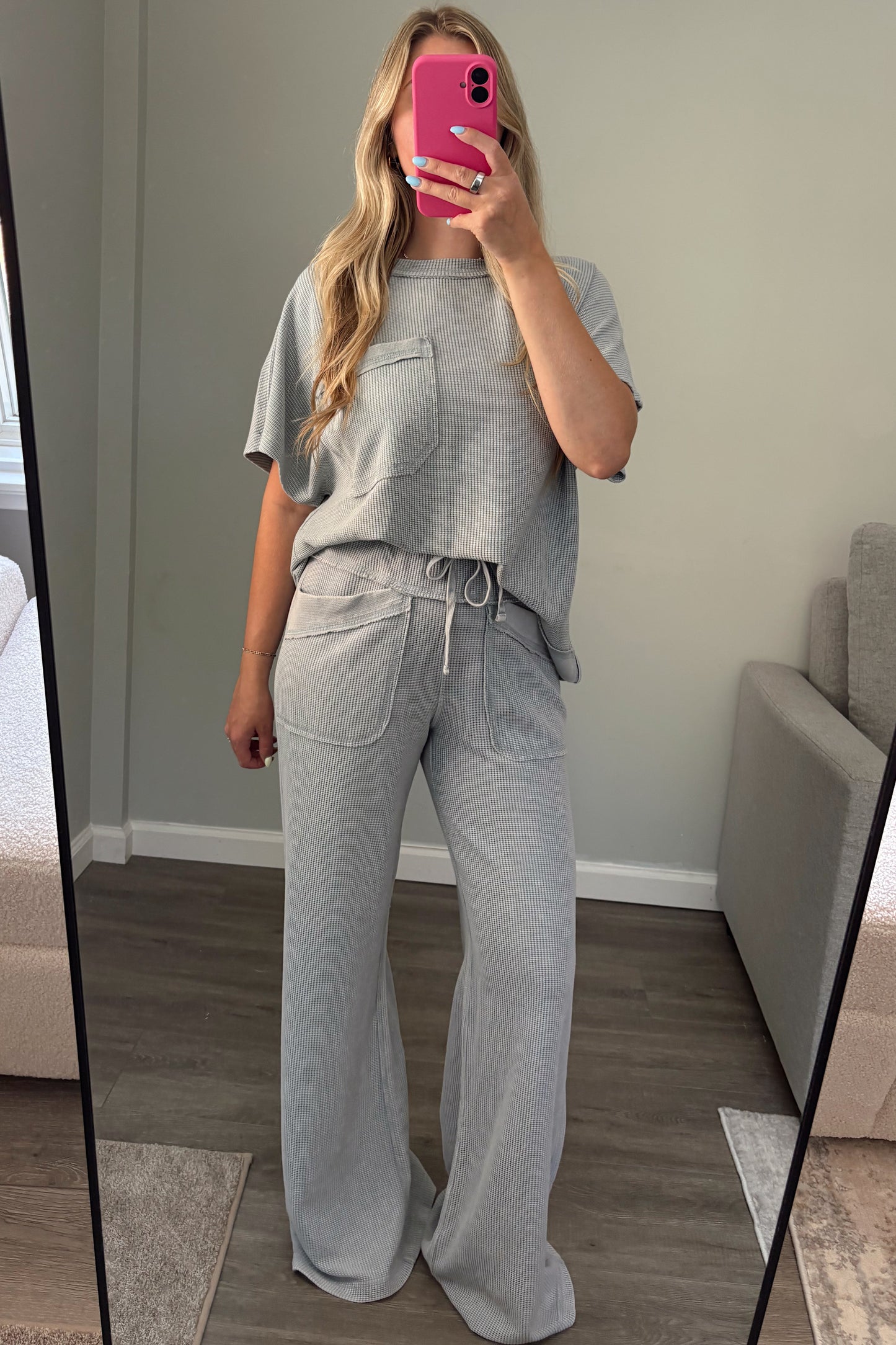 A woman takes a mirror selfie in a room wearing a light blue two-piece lounge set consisting of a short-sleeve top and matching wide-leg pants. She is holding a bright pink smartphone.