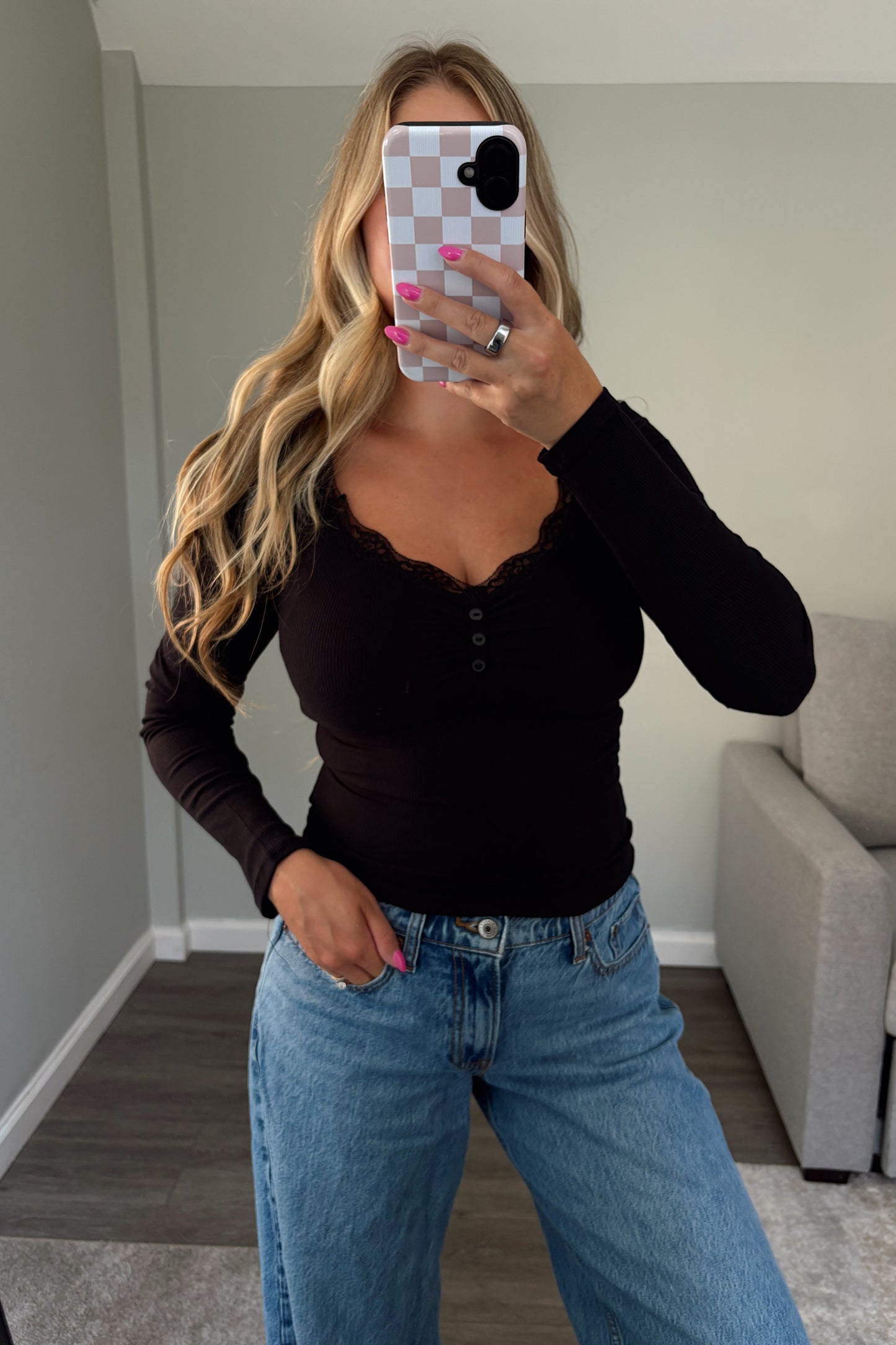 A woman takes a mirror selfie wearing a black long-sleeve henley top with a scalloped v-neckline and light-wash blue jeans in a modern room setting.