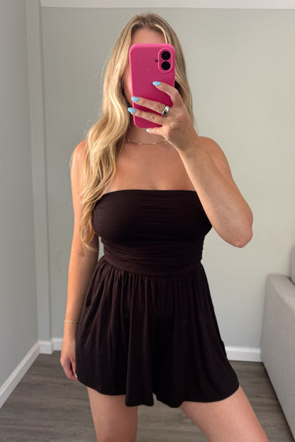 A woman takes a mirror selfie in a room wearing a strapless black romper. The background shows a gray wall, wood-look flooring, and a light gray sofa.