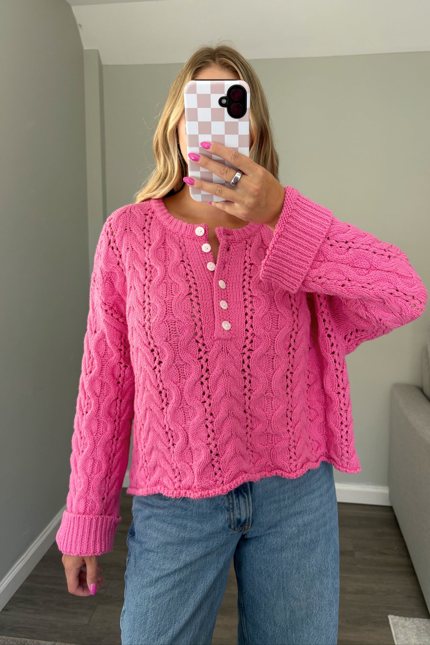 A woman takes a mirror selfie wearing a bright pink cable-knit cropped sweater with a buttoned henley neckline and blue jeans in a home setting.