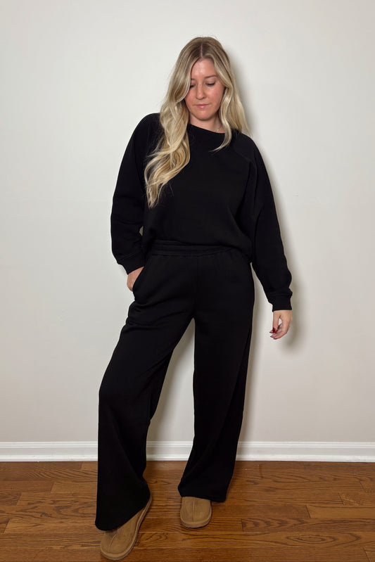 Person wearing a black lounge set standing against a plain wall.