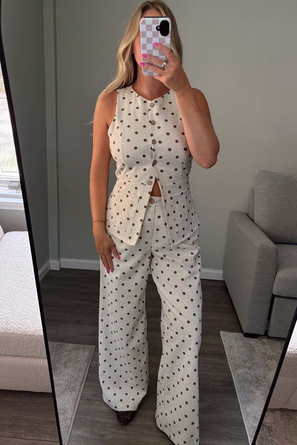 A woman takes a mirror selfie in a room showing a two-piece set with a white base and brown polka dots. The set includes a sleeveless button-front top with a small waist cutout and matching wide-leg pants.